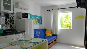 A compact living space with a kitchenette, a dining area with a table set for two, a blue sofa with cushions, and a window with a view of greenery.