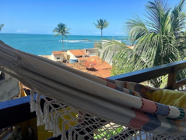 Una hamaca en un balcón con vista a una playa con palmeras y un cielo azul despejado.