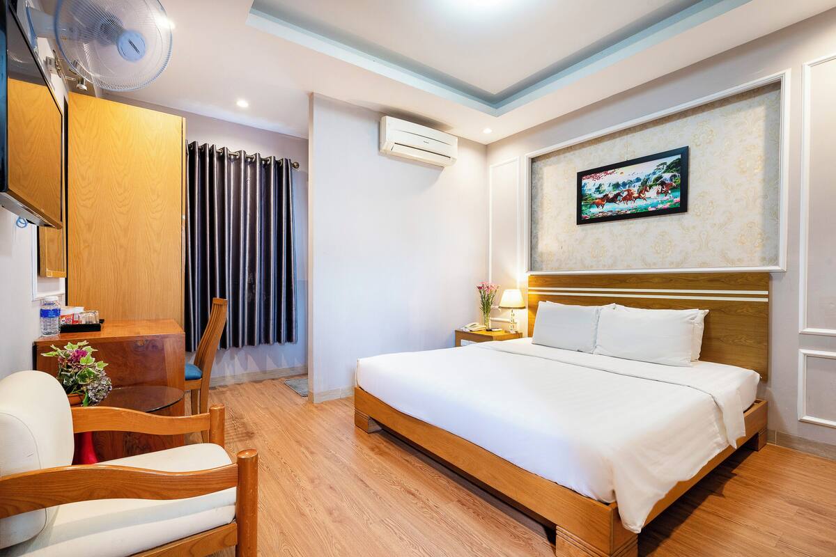 Une chambre d’hôtel avec un lit en bois, un bureau avec une chaise, un ventilateur sur pied, un appareil de climatisation et un tableau encadré au mur.