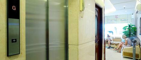 Un ascensor con pantalla digital y un teléfono a su lado. Dos personas están sentadas en un sofá al fondo.