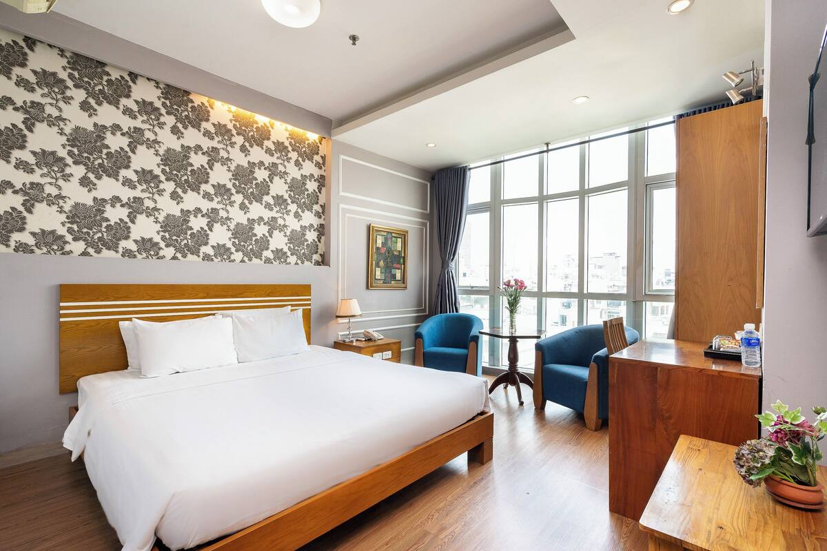 Une chambre d’hôtel avec un grand lit, un bureau, deux chaises, une petite table et une grande fenêtre donnant sur la ville.