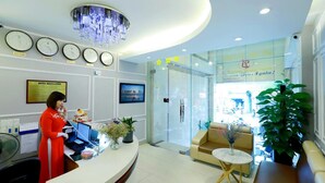 A hotel reception area with a receptionist, a clock displaying time in multiple cities, a chandelier, and a glass door leading to the lobby. - Lucky Star Hotel 266 De Tham (Ho Chi Minh City)