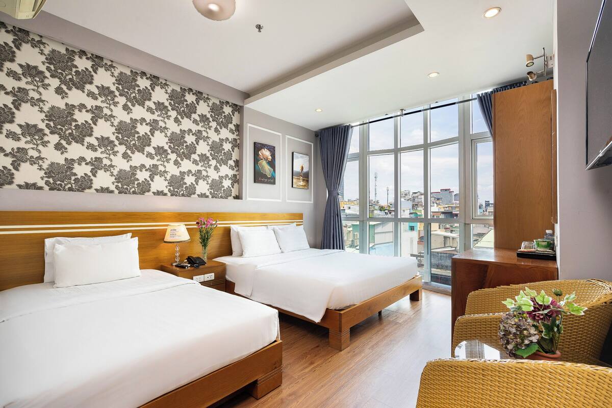 Une chambre d’hôtel avec deux lits, une grande fenêtre donnant sur la ville, une tête de lit en bois et un fauteuil en osier.
