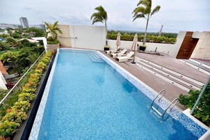 A rooftop swimming pool with clear blue water, surrounded by lounge chairs and umbrellas, with a view of the cityscape in the background.