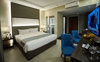 A hotel room with a bed, two blue armchairs, a desk with a bottle of wine and glasses, a TV displaying a news channel, and a bathroom visible through a glass partition.