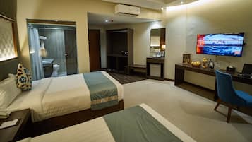 A hotel room with two beds, a desk with a computer, a TV displaying 'Tropical Storm', and a bathroom with a shower and toilet.