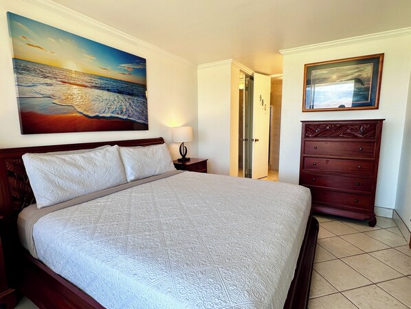 A hotel room with a large bed, a wooden dresser, a painting of a beach scene, and a door leading to a bathroom.