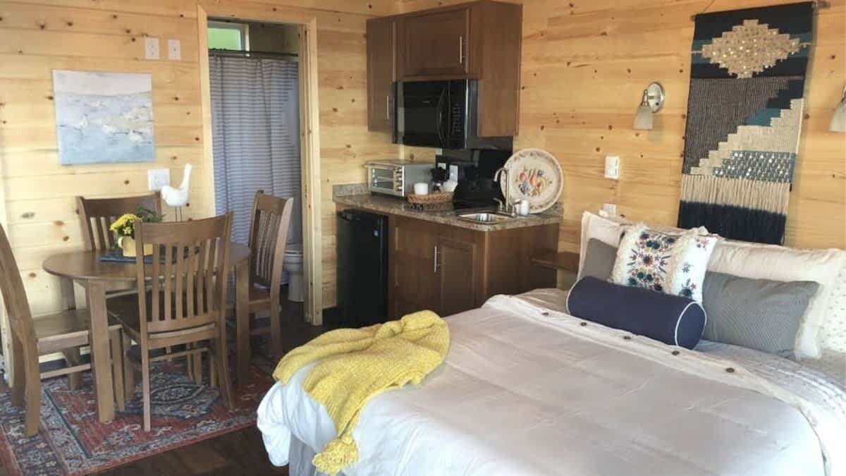 A cozy cabin-style room with a bed, a dining area with chairs, a kitchen with a microwave, and a decorative wall hanging.