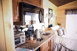 A compact kitchen with a microwave, toaster, and sink. There is a bed with pillows and a plant in the corner.