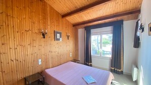 A room with a wooden wall, a bed, a small table, a window with curtains, and a radiator. - Charming Renovated House with Garden, 300m from Beach, WiFi Included (Bretignolles-sur-Mer)