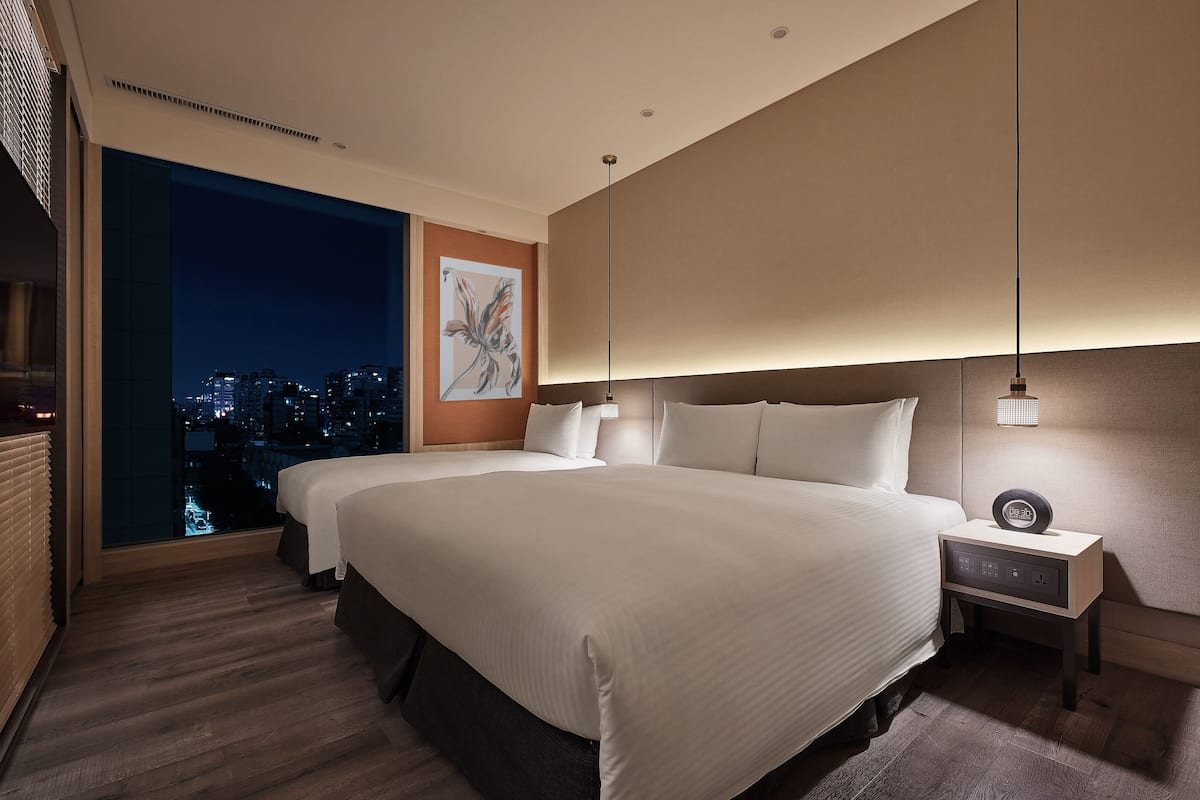 A modern hotel room with a large bed, a nightstand, and a city view through a large window.