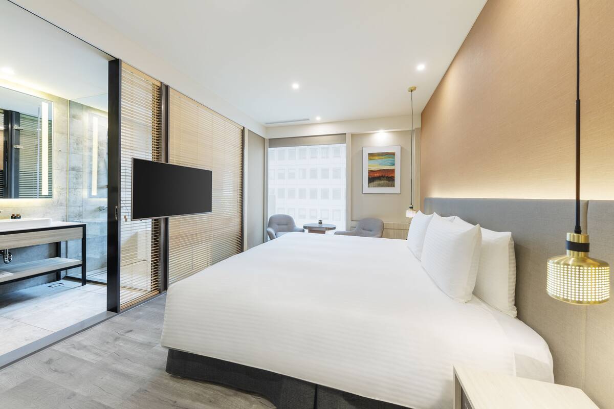A modern hotel room with a large bed, a TV, a desk with a chair, and a window with blinds.
