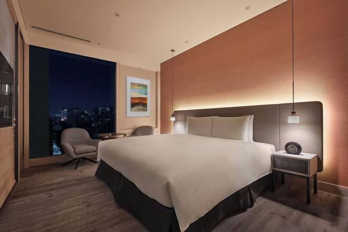 A modern hotel room with a large bed, a nightstand, a chair, and a large window with a city view.