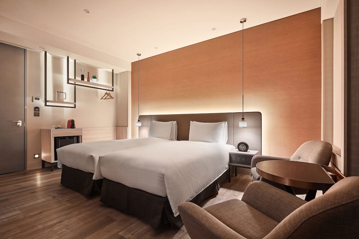A hotel room with two beds, a desk, a chair, a small table, and a wall-mounted shelf with decorative items.