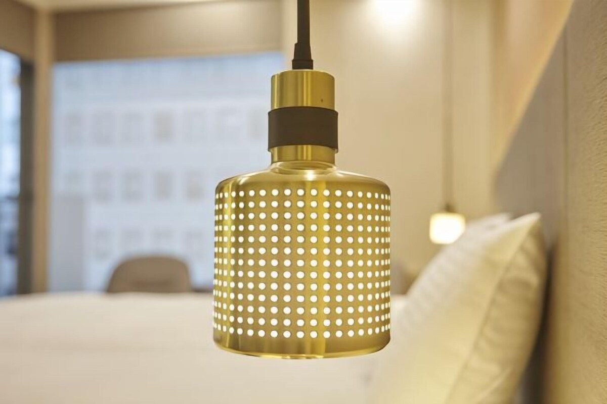 A pendant light fixture with a cylindrical shape and perforated pattern, hanging from a ceiling in a room with a bed and pillows.