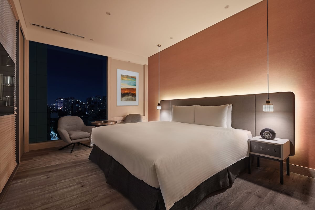 A modern hotel room with a large bed, a desk, a chair, and a view of the city at night.
