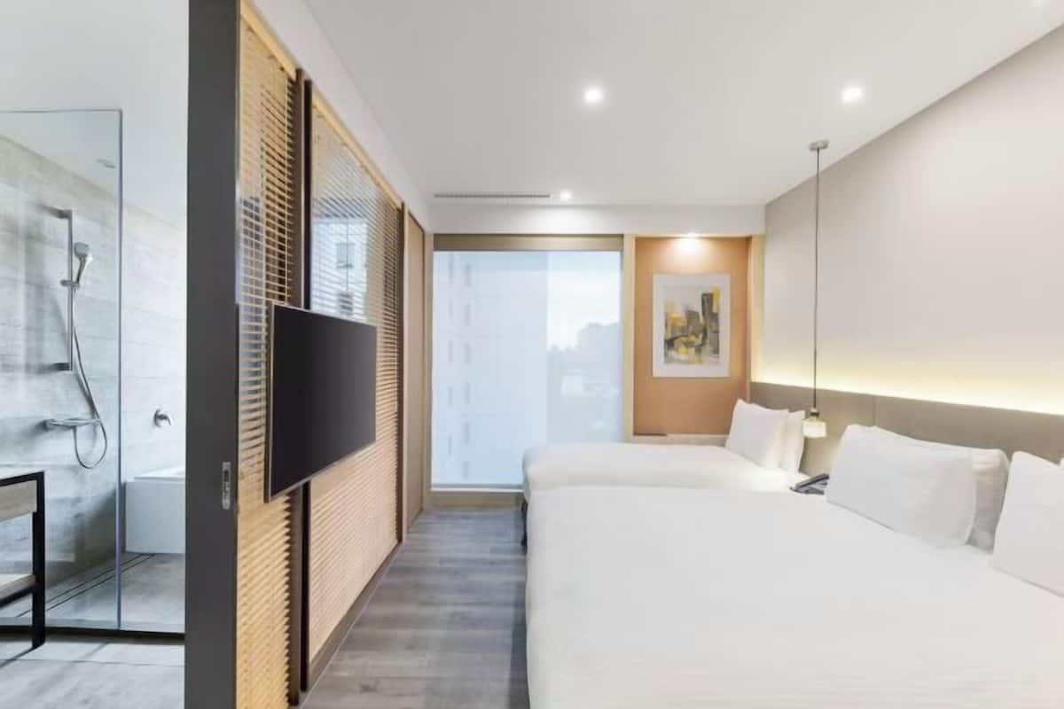 A hotel room with a bed, a TV, a shower, and a window with blinds.