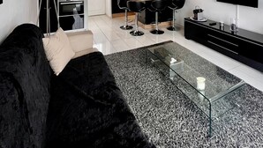 A modern living room with a black sofa, a glass coffee table, and a kitchen area in the background.