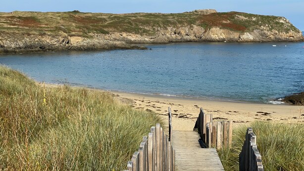 A wooden boardwalk leading to a sandy beach with a rocky cliff and calm sea.