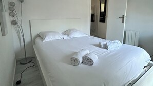 A neatly made bed with white linens and rolled towels, a bedside lamp, and a radiator.