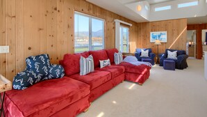 Ein modernes Wohnzimmer mit einem roten Ecksofa, blauen Sesseln und einem roten Hocker. Große Fenster bieten einen Ausblick ins Freie.