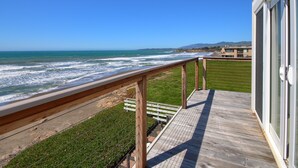 Una terraza de madera con barandilla da a una playa con olas, una zona verde y un edificio al fondo.