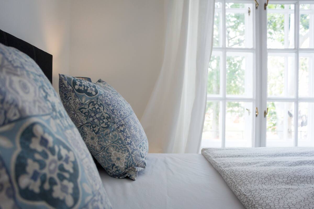 Une chambre à coucher avec un lit, des coussins à motifs et une fenêtre avec des rideaux.