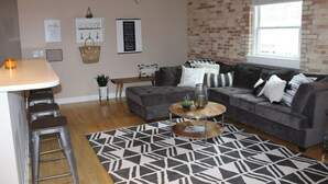 Ein modernes Wohnzimmer mit grauem Sofa, einem Couchtisch und einem gemusterten Teppich. Eine Ziegelwand und ein Fenster sind rechts zu sehen.