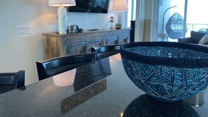 Un espace repas moderne avec une grande table noire, deux chaises, un vase décoratif, un téléviseur fixé au mur et une vue sur l’extérieur grâce à une grande fenêtre.