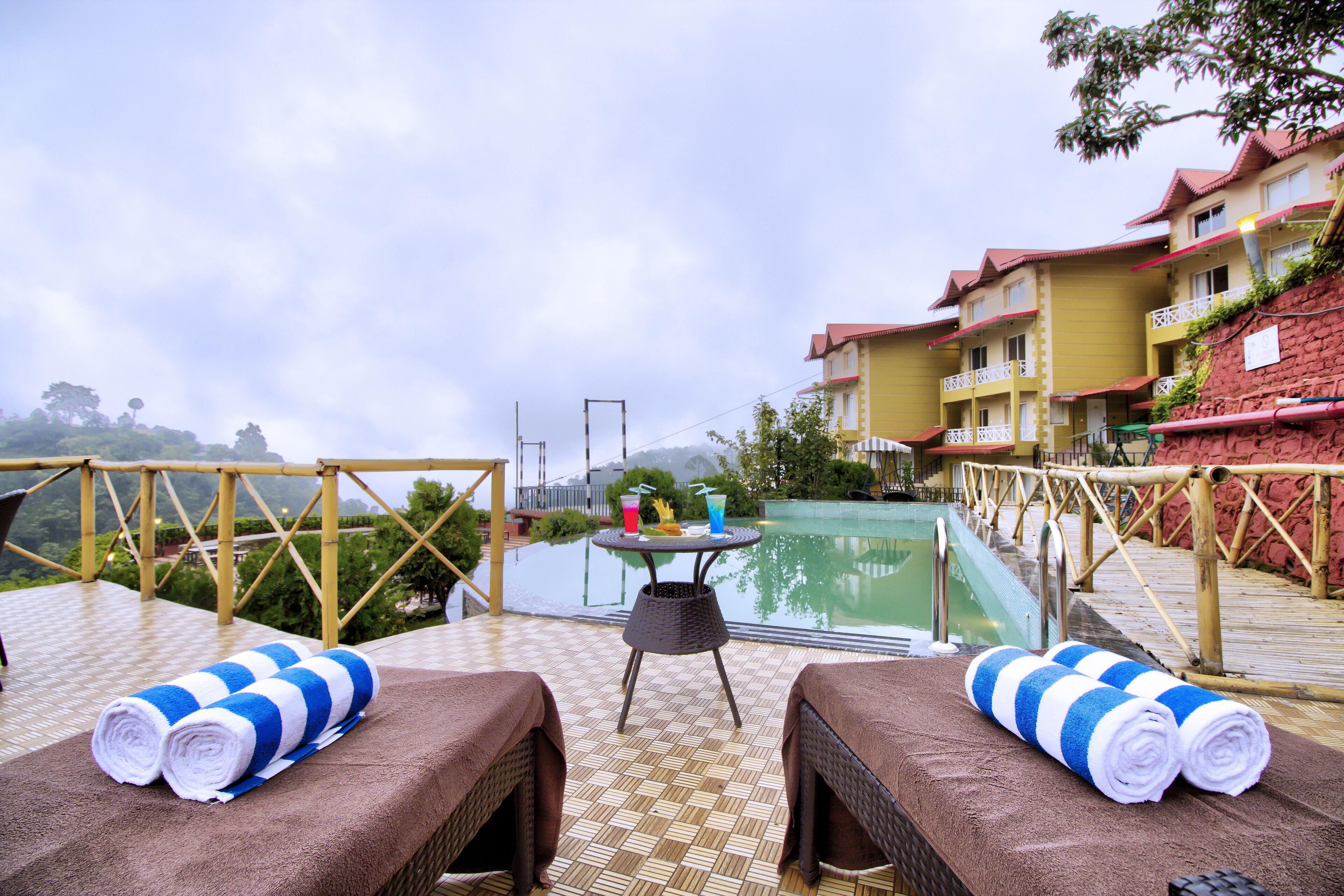 A poolside area with two sun loungers, a table with drinks, and a view of a pool and buildings in the background.