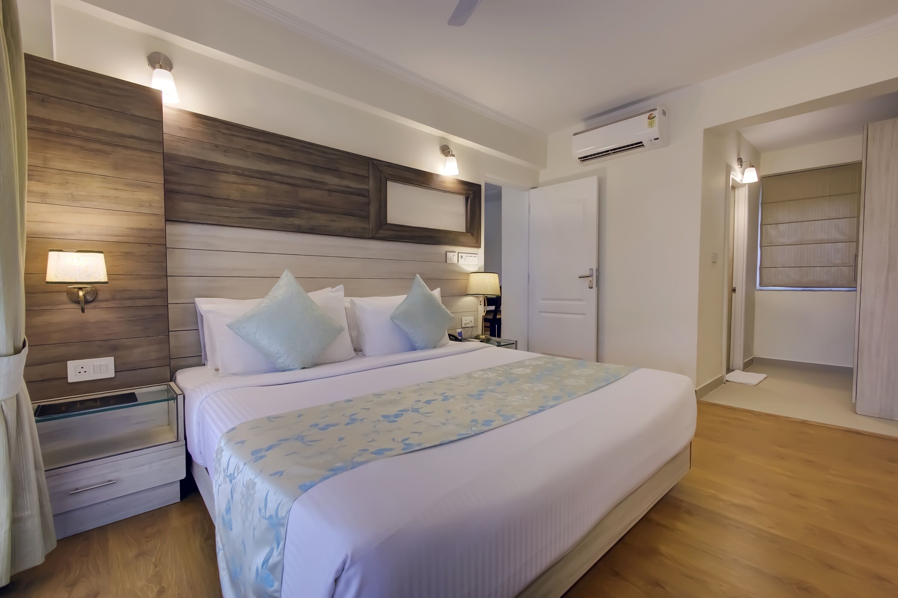 A hotel room with a large bed, wooden headboard, bedside lamps, a nightstand, and a window with curtains.