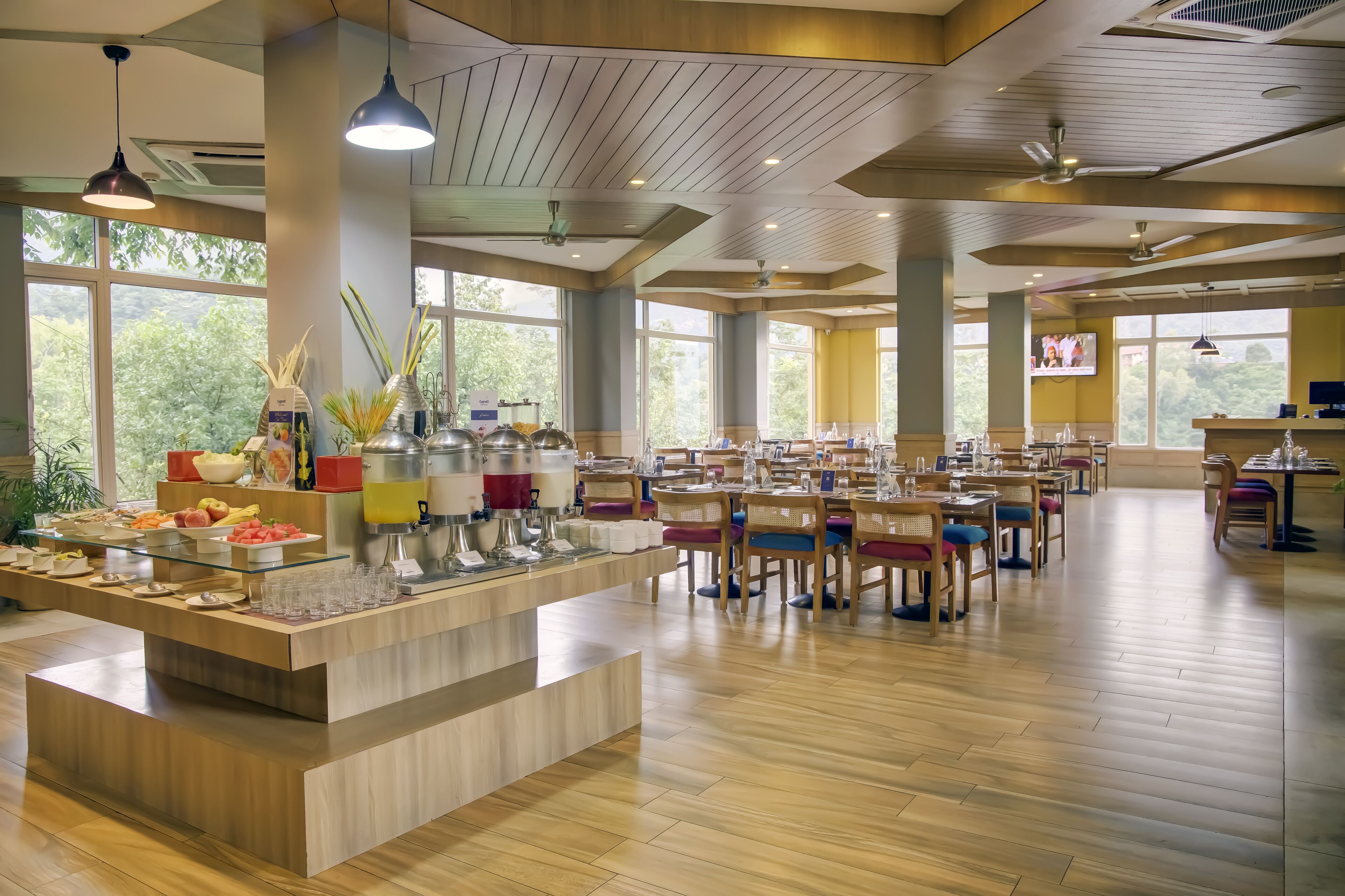 Un grand espace dédié à la salle à manger, avec de nombreuses tables et chaises, un buffet proposant une variété de plats et de grandes fenêtres laissant entrer la lumière naturelle.