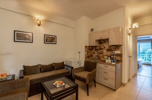 Un espacio habitable compacto con sofá, sillón y mesa de centro, una kitchenette con armarios de madera, y un balcón con barandilla.
