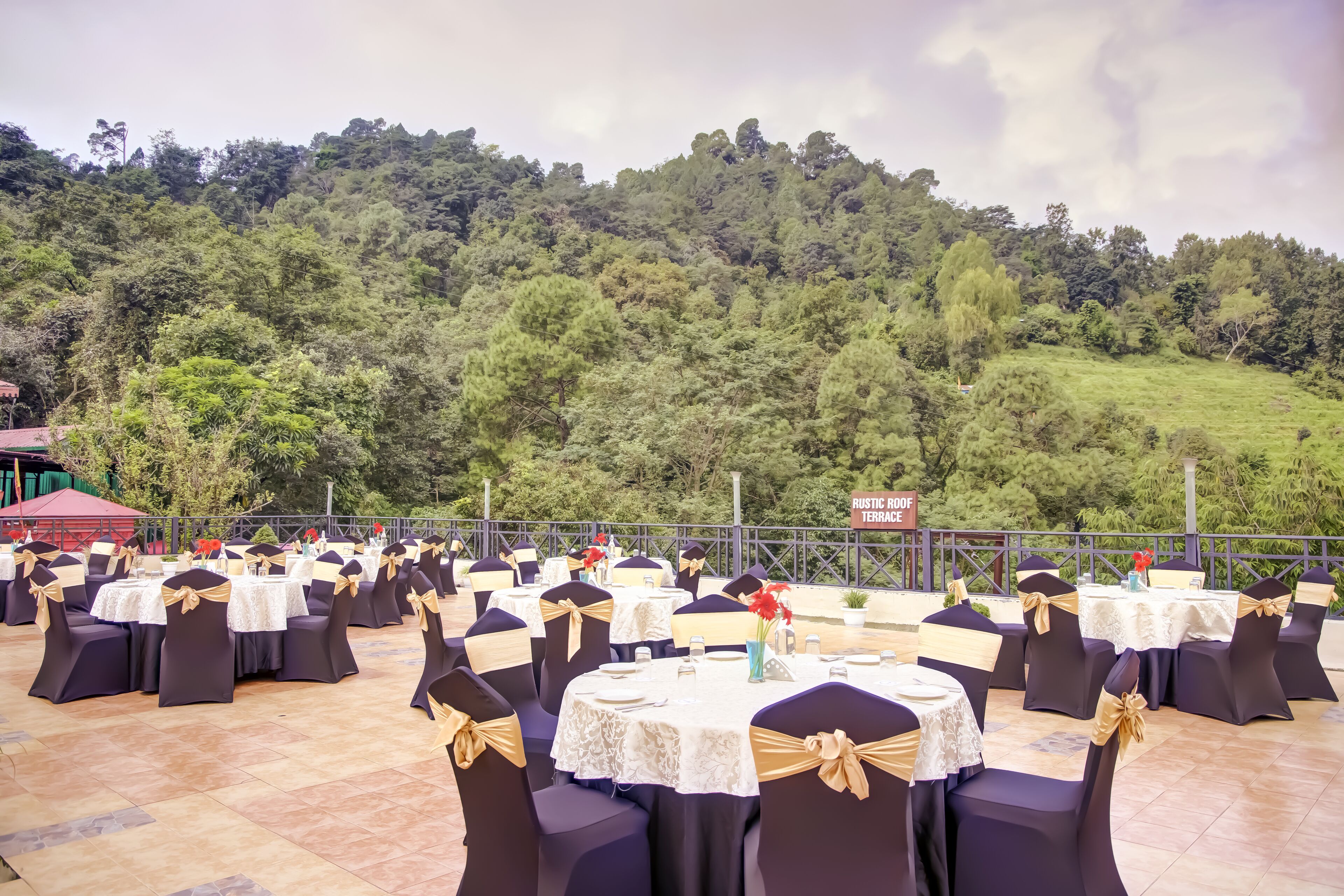 An outdoor event space with round tables and chairs, set against a backdrop of a lush green hillside.