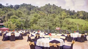 An outdoor event space with round tables and chairs, set against a backdrop of a lush green hillside.