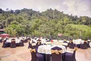 An outdoor event space with round tables and chairs, set against a backdrop of a lush green hillside.