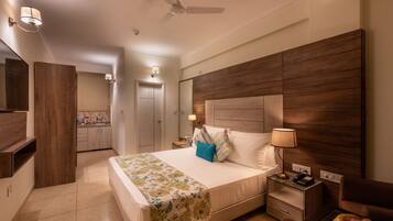 Quarto de hotel com cama grande, cabeceira de madeira, mesa de cabeceira com abajur e uma pequena mesa com frutas.