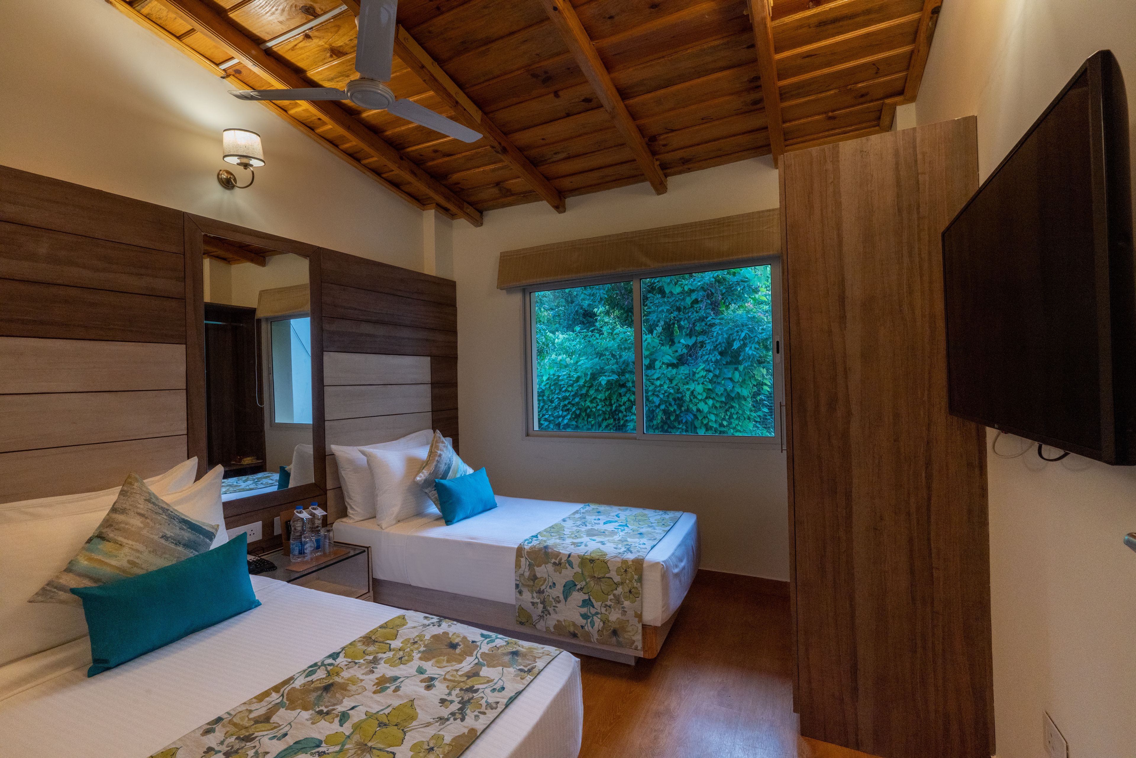 A room with two beds, a ceiling fan, a large window with a view of trees, and a mounted TV.