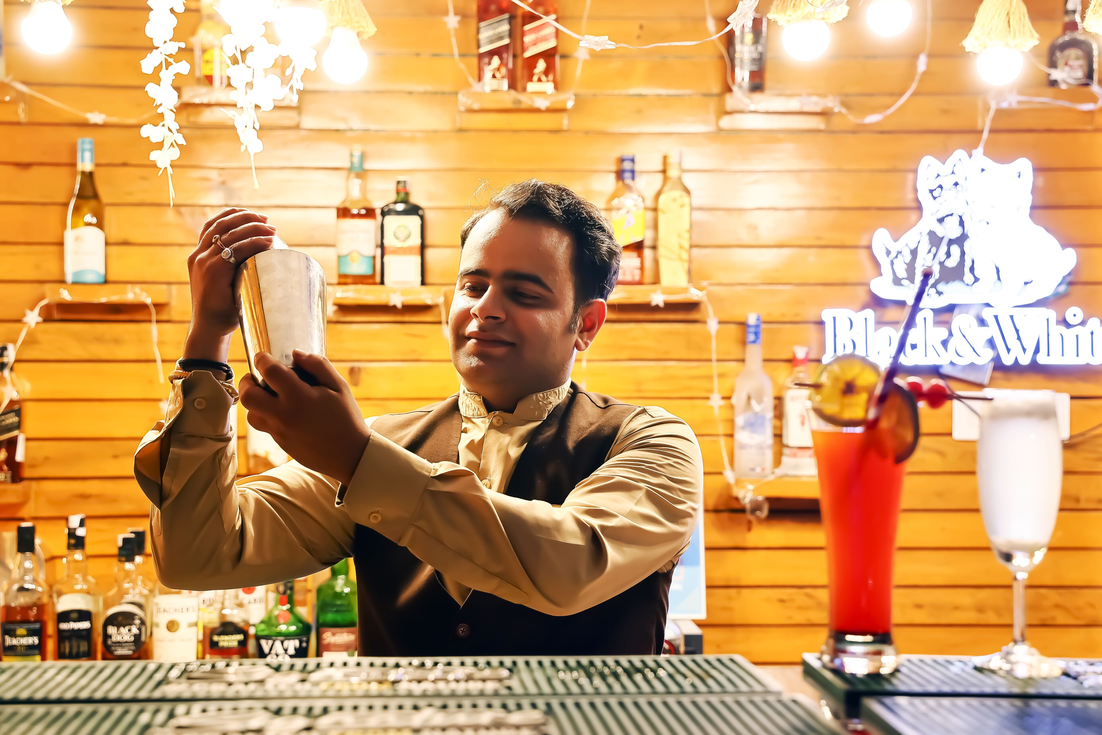 A bartender preparing a drink behind a bar with various liquor bottles and a neon sign in the background.