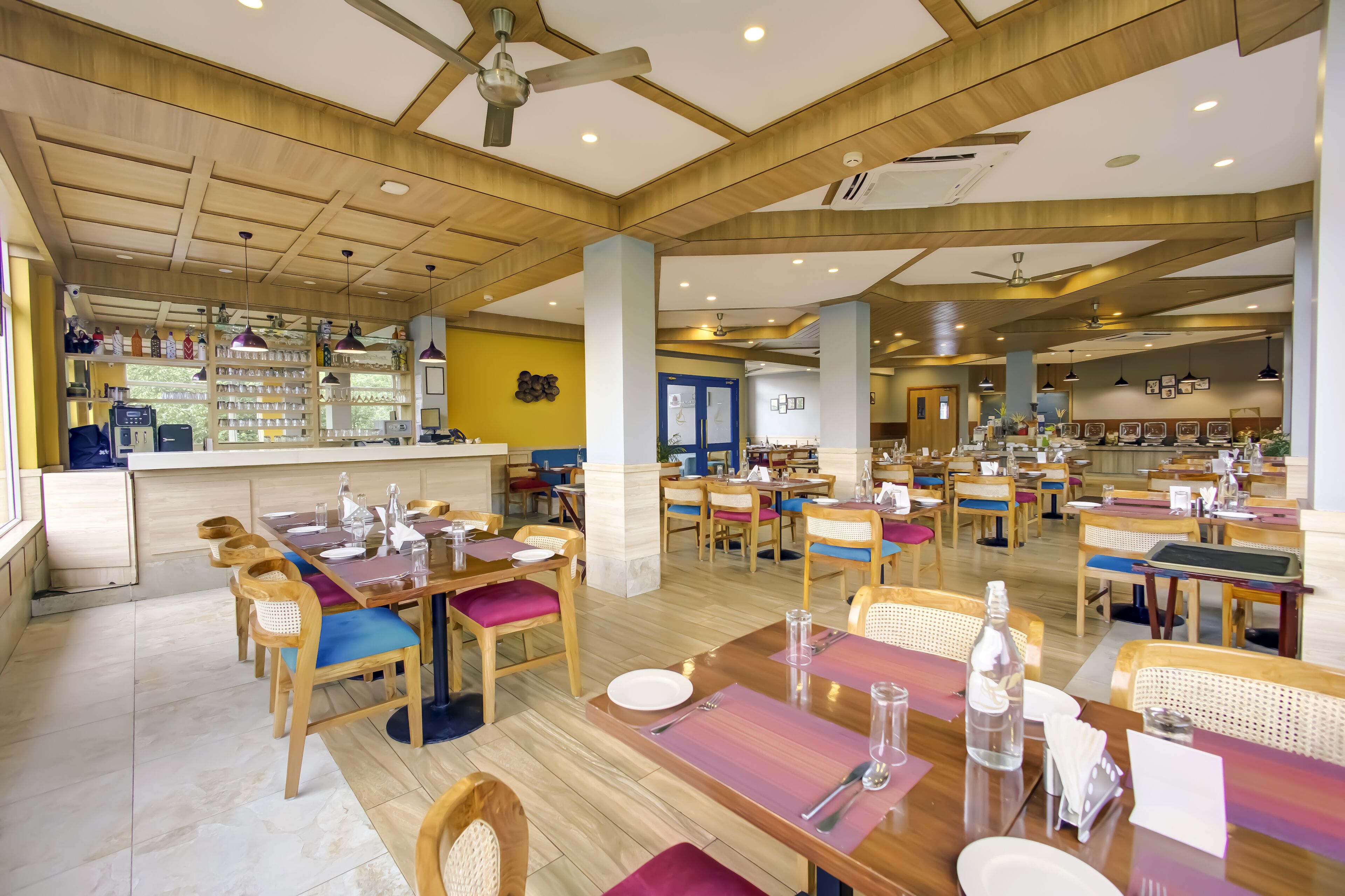 A spacious restaurant with wooden tables and chairs, a bar area, and a modern ceiling design.