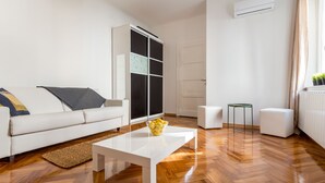 A modern living room with a white sofa, a white coffee table with a bowl of fruit, a small side table, and a radiator near a window with curtains. - Downtown Zagreb Apartments (Zagreb)