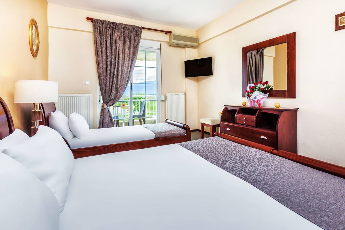 Habitación de hotel con una cama grande, una mesita de noche con lámpara, un aparador, un espejo y una ventana con cortinas.
