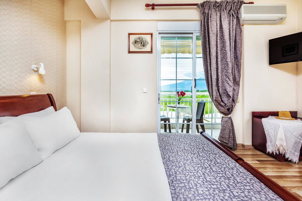 Habitación de hotel con cama, televisor, ventana con cortina y vista a un balcón con mobiliario exterior.