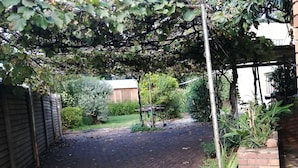 Ein gepflasterter Weg führt zu einem überdachten Gartenbereich, der auf einer Seite von einer Ziegelwand und auf der anderen von einem Metallzaun begrenzt wird.