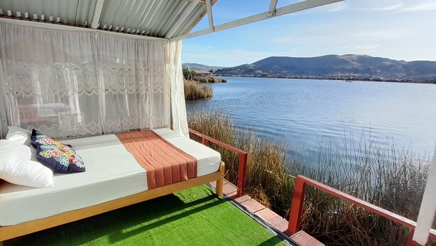 A balcony with a wooden bench, a white cushion, and a colorful pillow. The bench overlooks a calm lake with distant hills.