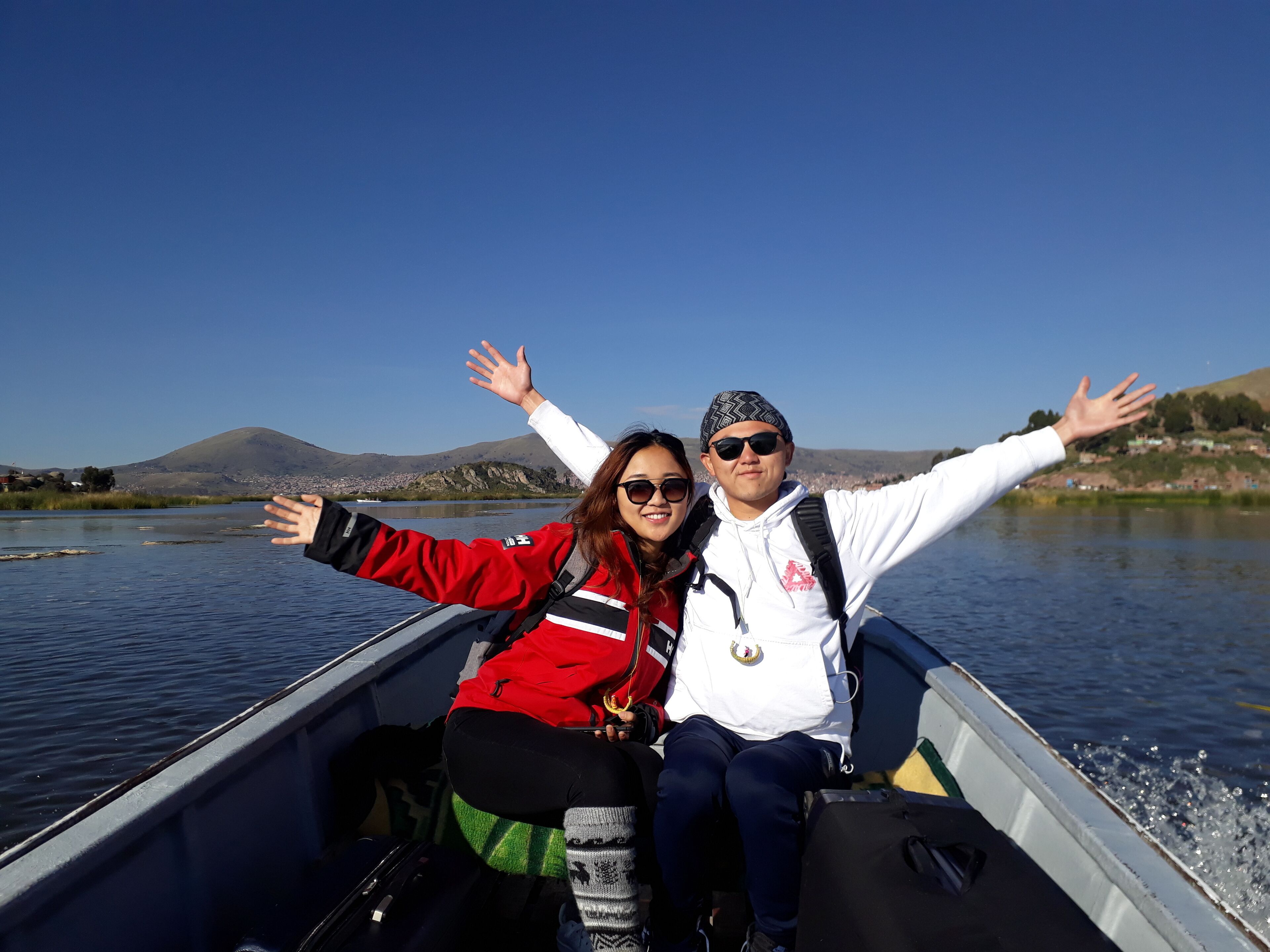 Two people in a boat with their arms outstretched, facing a calm lake with mountains in the background.