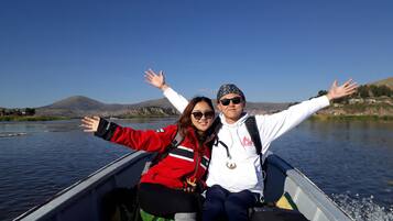 Two people in a boat with their arms outstretched, facing a calm lake with mountains in the background.