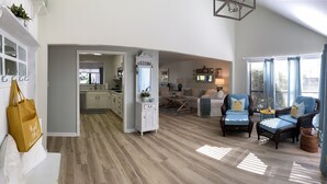 A spacious living area with a kitchen in the background, a white wall with a plant, and a large window with curtains. - Beautiful One-Story Cottage Just Minutes From White Sand Beach, with Golf Cart!  (Destin)