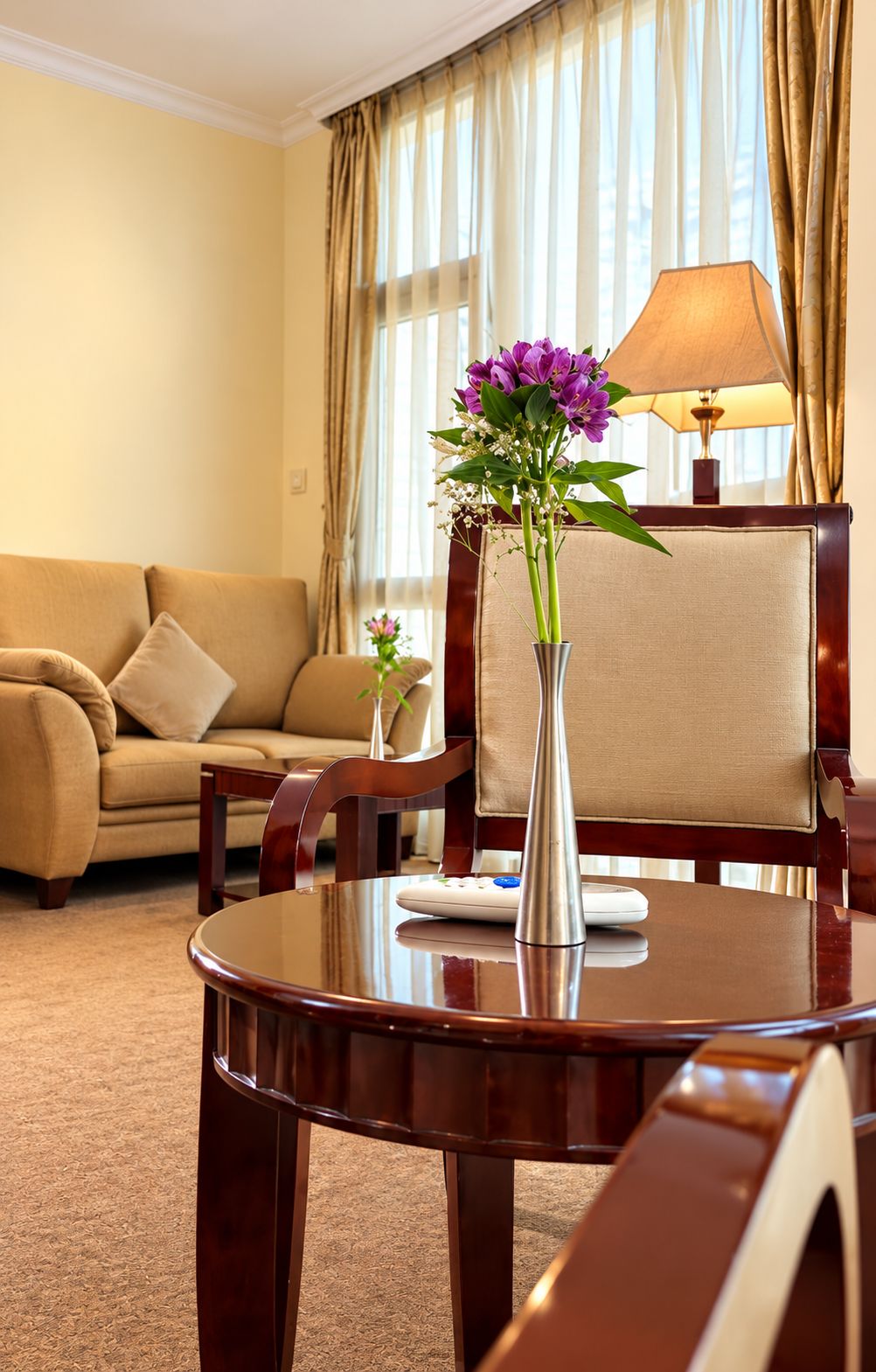 Een hotelkamer met een ronde tafel, een vaas met bloemen, een bank en een lamp.