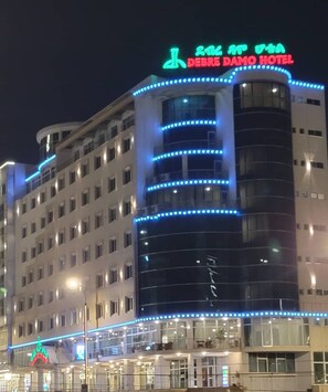 Um hotel com vários andares, iluminado por luzes azuis, e uma placa de neon indicando ser o Hotel Debre Damo.